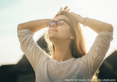 Woman with eyes closed and hands on head, smiling and looking up at the sun.